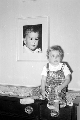 A black and white photograph of a young child standing next to a framed portrait of another young child. The child is wearing...