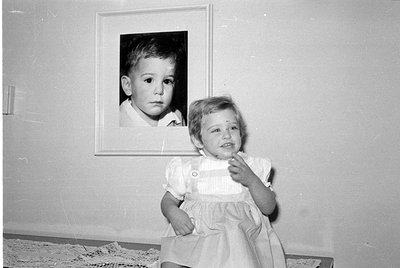 A black and white photograph oriented upside down. A young child in a light-colored dress is positioned next to a framed port...
