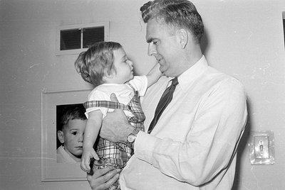 A black and white photograph of a man in a white dress shirt and tie holding a small child in plaid overalls. A framed portra...