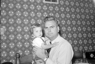 A black and white photograph of a man and a young child. The man wears a light-colored button-down shirt and a dark tie, hold...