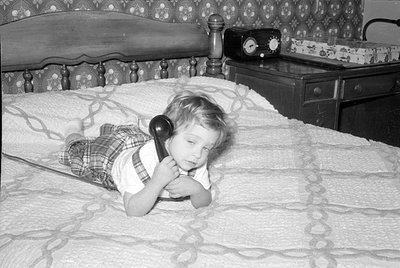 A young child in plaid trousers and a white shirt lies on a patterned bedspread, holding a black rotary phone receiver to the...