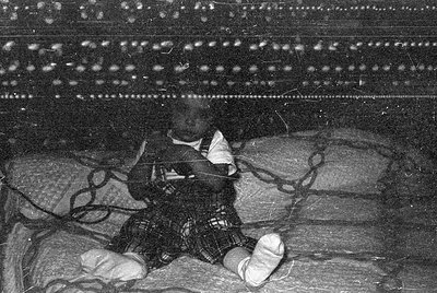 A black and white photograph of an infant sitting on a patterned fabric surface. The child wears a light-colored shirt and da...