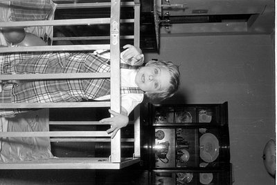 A black and white photograph of a young child wearing a plaid dress and white collar, looking through the bars of a wooden cr...