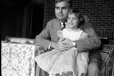 A black and white photograph of a man in a suit and a young girl in a light-colored dress with a white collar. They are seate...