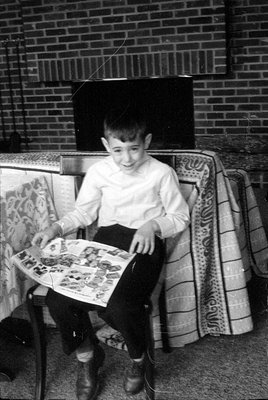 A black and white photograph of a young boy sitting on a chair and holding an open magazine. He is positioned in front of a b...