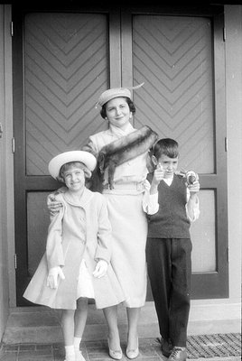 A black and white studio-style portrait of a woman and two children standing before a pair of dark doors with chevron-pattern...