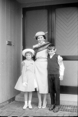 A black and white photograph of a woman and two children standing outdoors on a tiled porch. The woman wears a light-colored ...