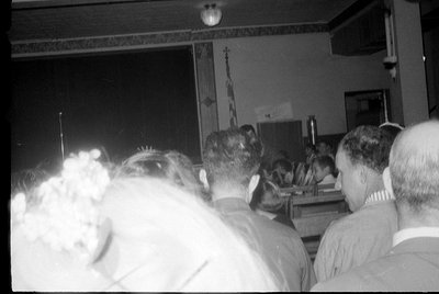 Black and white photograph showing the backs of several people seated in a room, facing a dark stage or screen. A few individ...
