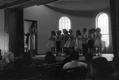 A group of children in formal attire stand in a line on a stage, some with hands clasped. A woman in a patterned dress stands...