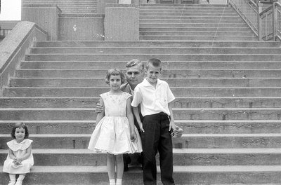 A black and white photograph of a man and two children sitting on wide concrete stairs, with a third small child sitting on a...