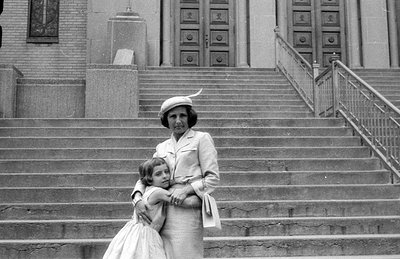A woman in a light-colored suit and a small hat stands on wide stone stairs, holding a young girl in a light-colored dress. B...