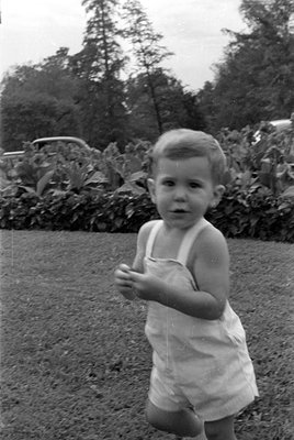 A black and white photograph of a young child wearing a light-colored sleeveless romper. The child is positioned in front of ...