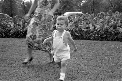 A toddler in white overalls and socks walks across a grassy lawn. In the background, a person in a patterned dress is capture...