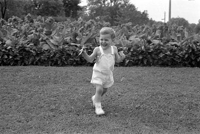 A black and white photograph of a young child wearing light-colored overalls and white socks, running across a grassy lawn. T...