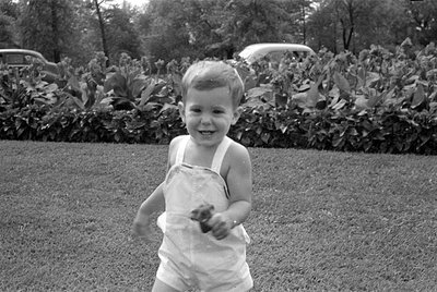 A black and white photograph, oriented upside down, showing a young child standing outdoors. The child is wearing a light-col...