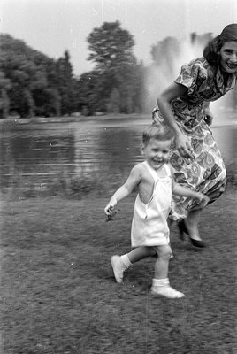 A black and white photograph of a young child in white overalls and white shoes running on grass. Behind the child, a woman i...