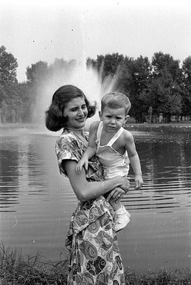 A black and white photograph of a woman in a patterned dress holding a small child in a white romper. They are standing in fr...
