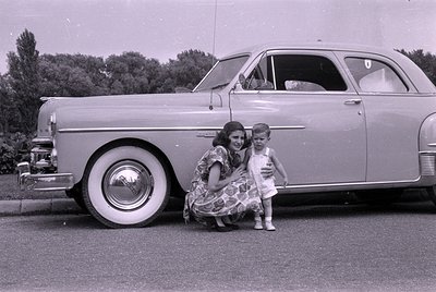 A woman in a patterned dress and a small child in a white outfit pose beside a light-colored vintage sedan with whitewall tir...