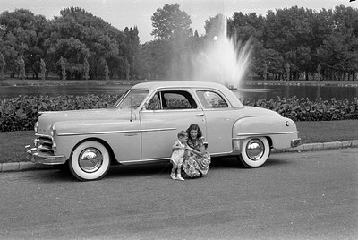 A side-profile view of a light-colored 1940s-era sedan parked on a paved road. A woman in a patterned dress and a small child...