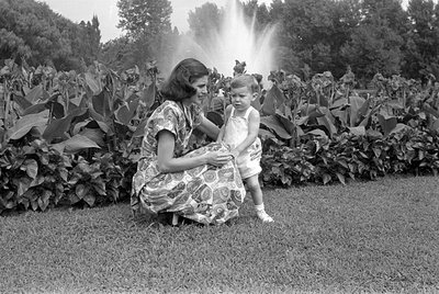 A black and white photograph of a woman and a small child in a garden. The woman, wearing a patterned dress, is crouching nex...
