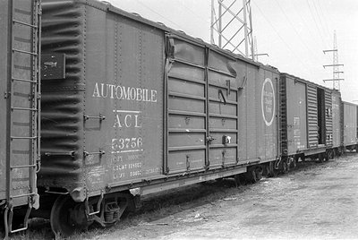A black and white photograph of a line of railway freight cars. The closest car is marked 'AUTOMOBILE' and 'ACL 53756'. High-...