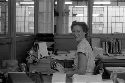 A woman in a striped short-sleeved shirt sits at a desk with a typewriter, looking toward the camera. The office setting incl...
