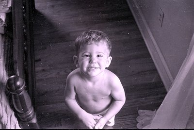 A black and white photograph of a young child sitting on a wooden floor. The child is shirtless and looking toward the camera...