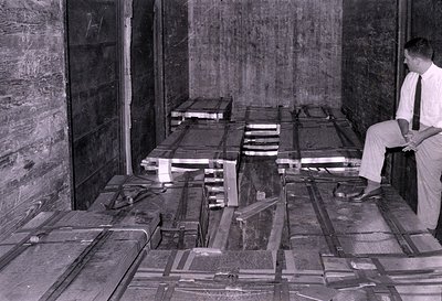 A black and white photograph showing the interior of a wooden cargo container or room. Several large, rectangular crates wrap...