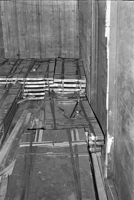 Black and white photograph of stacked metal sheets or slabs secured with straps. The materials are stored in a concrete room ...