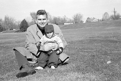 A black and white photograph of a man and a small child sitting on a grassy slope. The man wears a light-colored overcoat and...