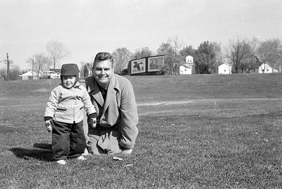 A man and a small child pose in a grassy field. The man is kneeling and wearing a light-colored overcoat, while the child sta...