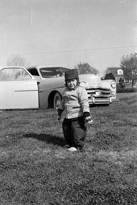 A young child wearing a dark hat, light-colored jacket, and dark trousers stands in a grassy field. Behind the child is a lig...