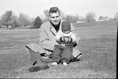 A man in a light-colored overcoat sits on a grassy field, holding a small child. The child wears a dark cap, a light jacket, ...