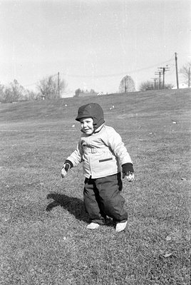 A black and white photograph of a small child standing in a grassy field. The child is wearing a light-colored winter jacket,...