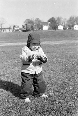 A black and white photograph of a small child standing in a grassy field. The child wears a hooded jacket, dark trousers, and...