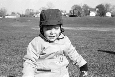 A black and white photograph of a young child sitting in a grassy field. The child wears a light-colored jacket with a belt a...