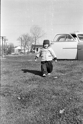 A young child wearing a dark hat, light-colored jacket, and dark trousers walks across a grassy yard. A light-colored vintage...