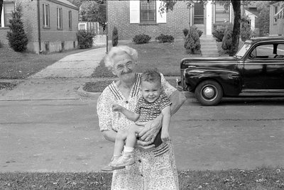 A black and white photograph of a woman in a floral print dress holding a small child. They are standing on a residential str...
