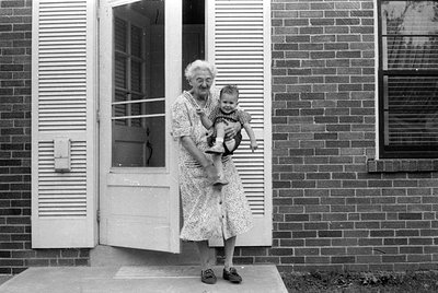 A black and white photograph of an older woman standing in a doorway of a brick house, holding a small child. The woman wears...