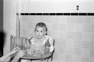 A black and white photograph of a toddler sitting in a high chair against a tiled wall. The child wears a ruffled bib and has...