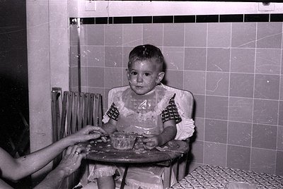 A black and white photograph of a young child sitting in a high chair with a plastic bib. The child has food on their face an...