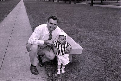 A black and white photograph of a man and a young child outdoors. The man, wearing a light-colored shirt and tie, is crouchin...