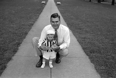 A black and white photograph of a man and a young child posing on a concrete sidewalk. The man wears a white button-down shir...