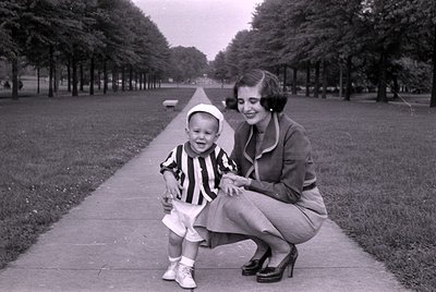 A black and white photograph of a woman and a young boy on a paved walkway in a park. The boy wears a striped shirt, white sh...