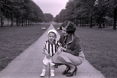 A woman and a young child pose on a concrete path lined with rows of trees. The child wears a striped shirt, white shorts, an...