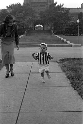 A black and white photograph of a young child in a striped shirt and white shorts walking on a concrete path. A woman in a sk...