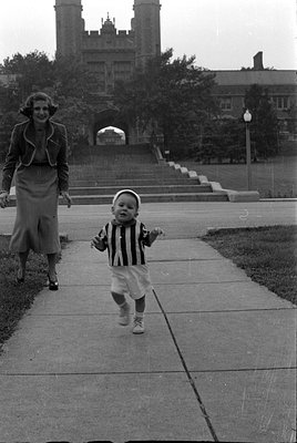 A black and white photograph of a toddler in a striped shirt and white shorts walking toward the camera on a concrete path. A...