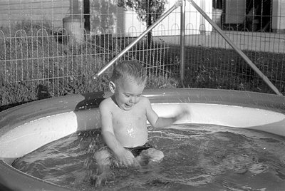 A black and white photograph of a young child sitting in a small, inflatable swimming pool. The child is splashing in the wat...