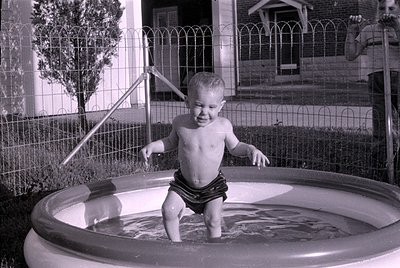 A black and white photograph of a shirtless young child standing in a small, circular inflatable pool. In the background, a w...