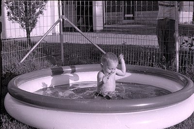 A young child sits in a small, circular inflatable swimming pool filled with water. In the background, a wire fence and a por...